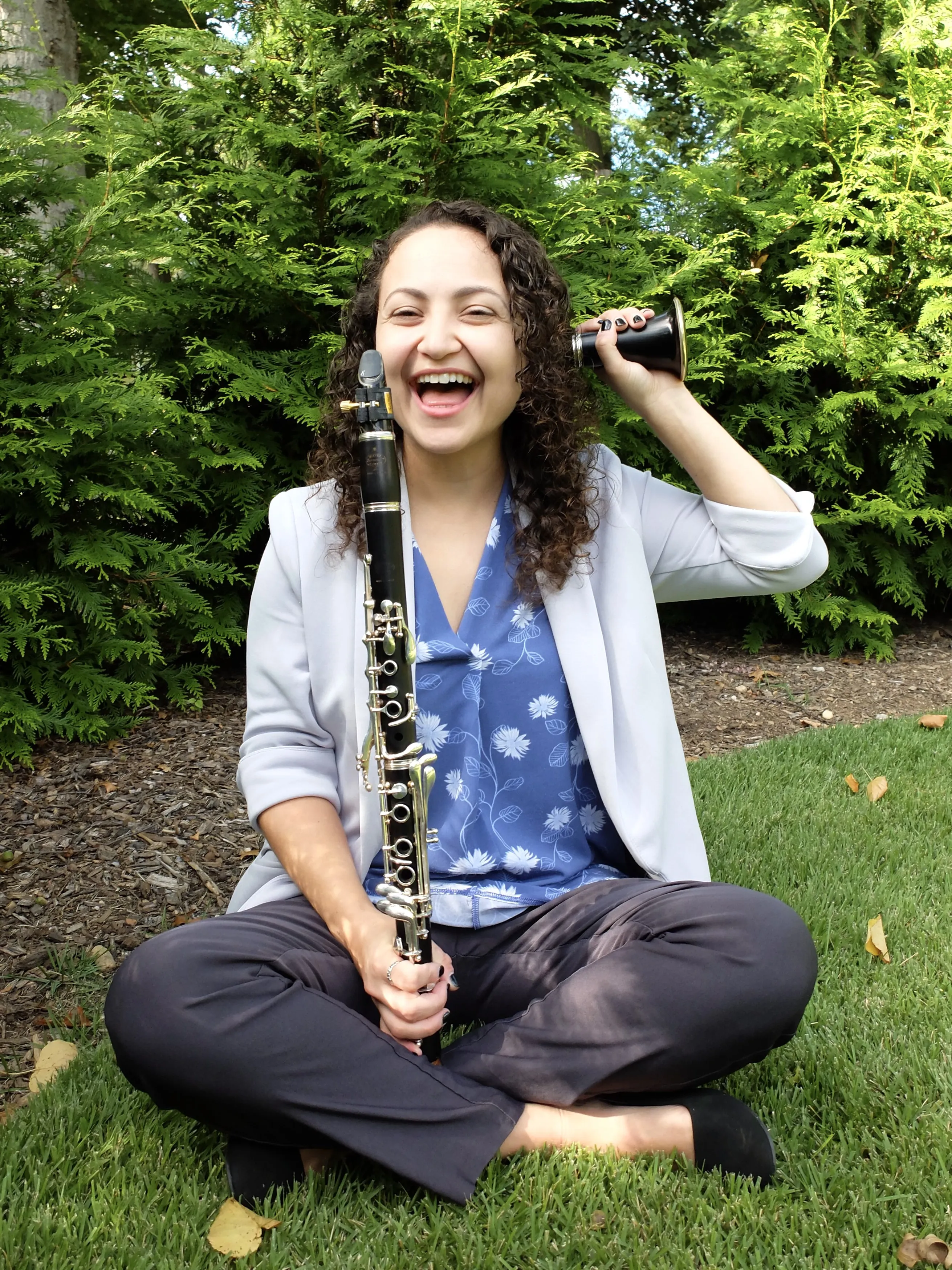 Valerie sitting criss-cross applesauce in the grass, giggling, while holding the detached bell of her clarinet to her right ear, and the rest of her clarinet upright in the other